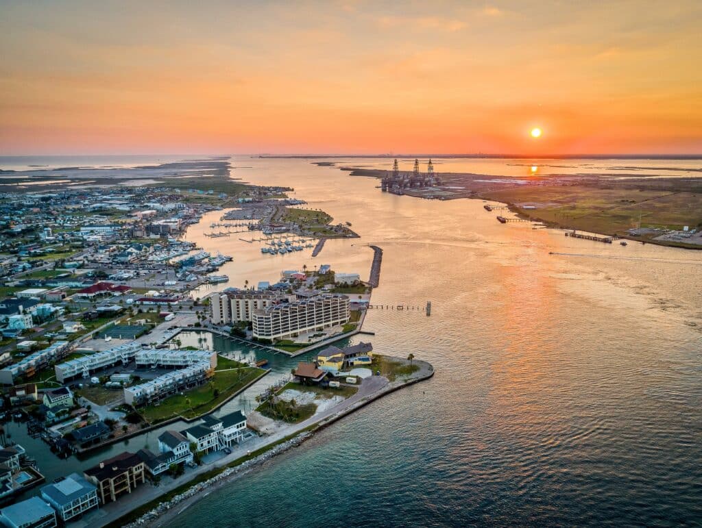 Aerial view of a coastal town during sunset with waterways and docks. The skyline features hotels and residential buildings along the water, reflecting the evening colors. This scene depicts the beauty of home construction in Corpus Christi.