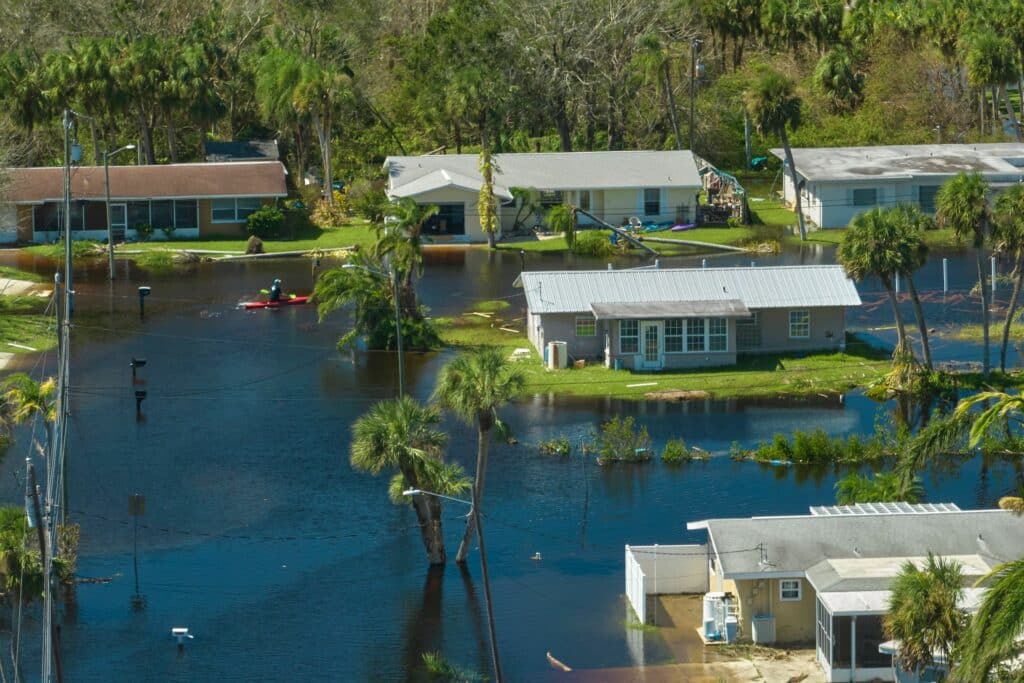 An aerial view of a flooded residential area, with homes partially submerged in water. A person is seen paddling a kayak through the flooded streets, surrounded by palm trees and debris, illustrating the impact of severe flooding.