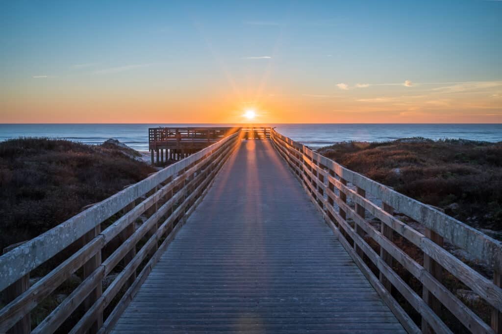A wooden walkway leading to the beach with a vibrant sunset on the horizon. The sun is partially visible, casting rays over the ocean waves and a grassy area on either side of the path. This image represents natural beauty in Southern Texas.