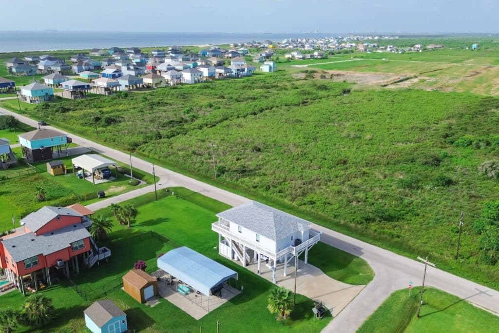 Aerial view of a coastal neighborhood with colorful houses along a road. Green fields are visible nearby, and a waterfront is seen in the background. The scene captures a mix of residential and natural landscapes.