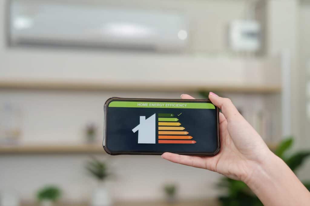 A person holding a smartphone displaying a graphic related to home energy efficiency, featuring a house icon and colored bars indicating efficiency levels. In the background, there are indoor plants and modern decor.