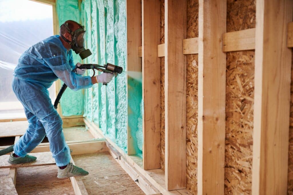 A worker in protective clothing applying insulation foam to a wooden wall frame in a construction site, with green insulation material visible.