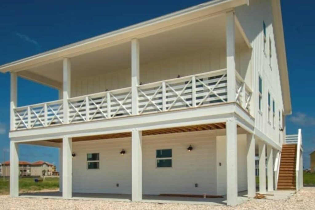 A modern two-story white house with a spacious balcony and wooden stairs leading up to it. The ground level features several windows and outdoor lighting. The surrounding area has a clear blue sky.