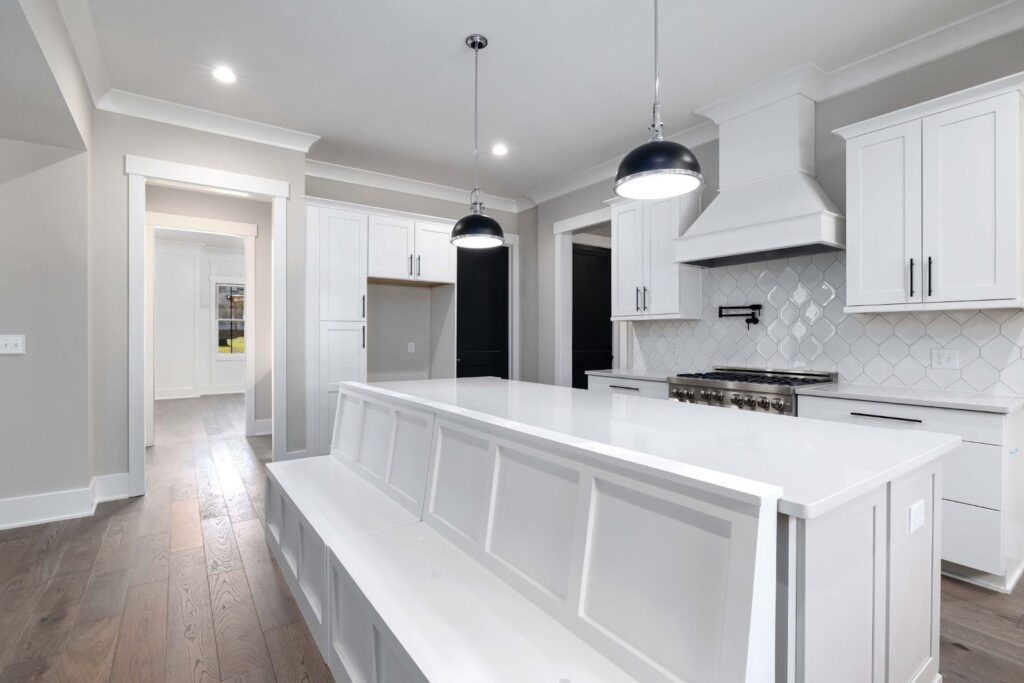 A modern kitchen featuring a large white island with seating, pendant lights, and stainless steel appliances. White cabinets and a tiled backsplash complement the wood flooring and light gray walls.