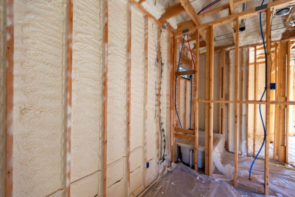 An interior construction site showing wooden framing with insulated walls covered in spray foam. Electrical wires are visible, and the floor is covered with protective plastic sheeting.
