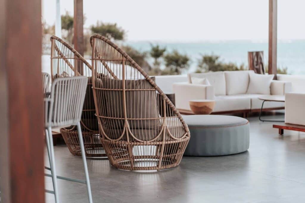 Two woven rattan chairs positioned near a blue ottoman in a contemporary lounge area, with light-colored seating and a view of the ocean visible through large windows.