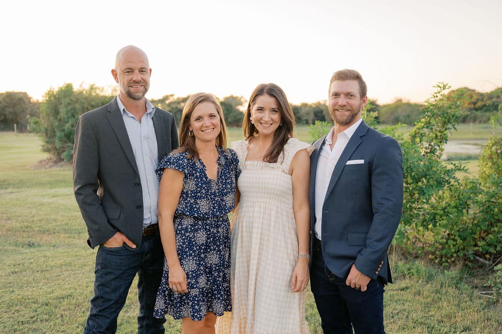cth fam Four adults posing outdoors in a grassy area during sunset. The first person on the left is wearing a suit jacket and jeans, the second is in a blue dress, the third is dressed in a white dress, and the last is in a blazer and shirt, all smiling at the camera.