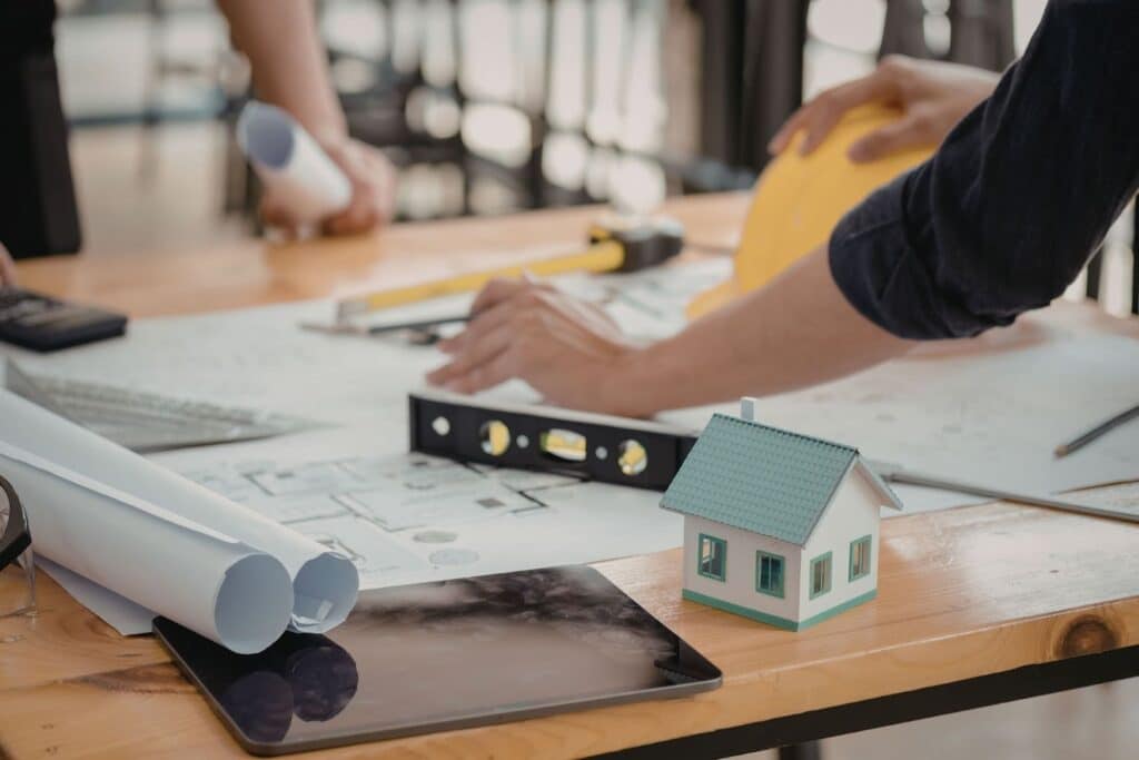 A workspace with blueprints and drafting tools on a wooden table. Hands are positioned over plans, with a small model house nearby. An electronic tablet is also visible, along with a level and measuring tools.