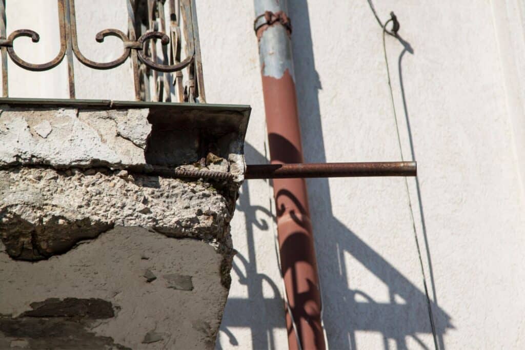 A close-up view of a weathered concrete balcony showing a large crack and exposed reinforcing rod. In the background, a rusty vertical pipe and a shadow cast by the balcony railing are visible on the wall.