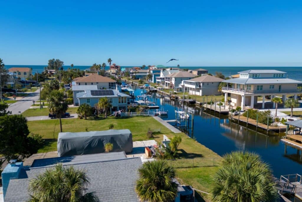 An aerial view of a coastal residential area featuring several houses and boat docks along a canal. The scene includes palm trees and a clear blue sky, with the ocean visible in the background.