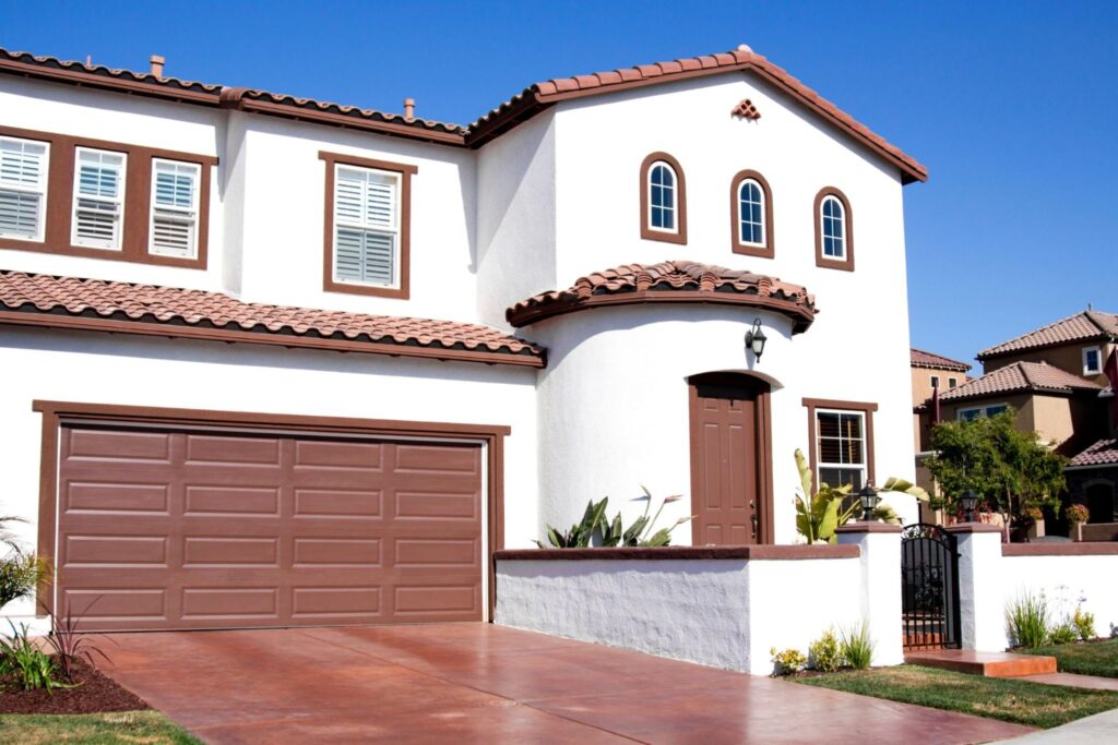 A modern two-story house with white stucco walls and a brown tiled roof. The front features arched windows, a garage door, and a decorative front door, surrounded by landscaping and a concrete driveway.